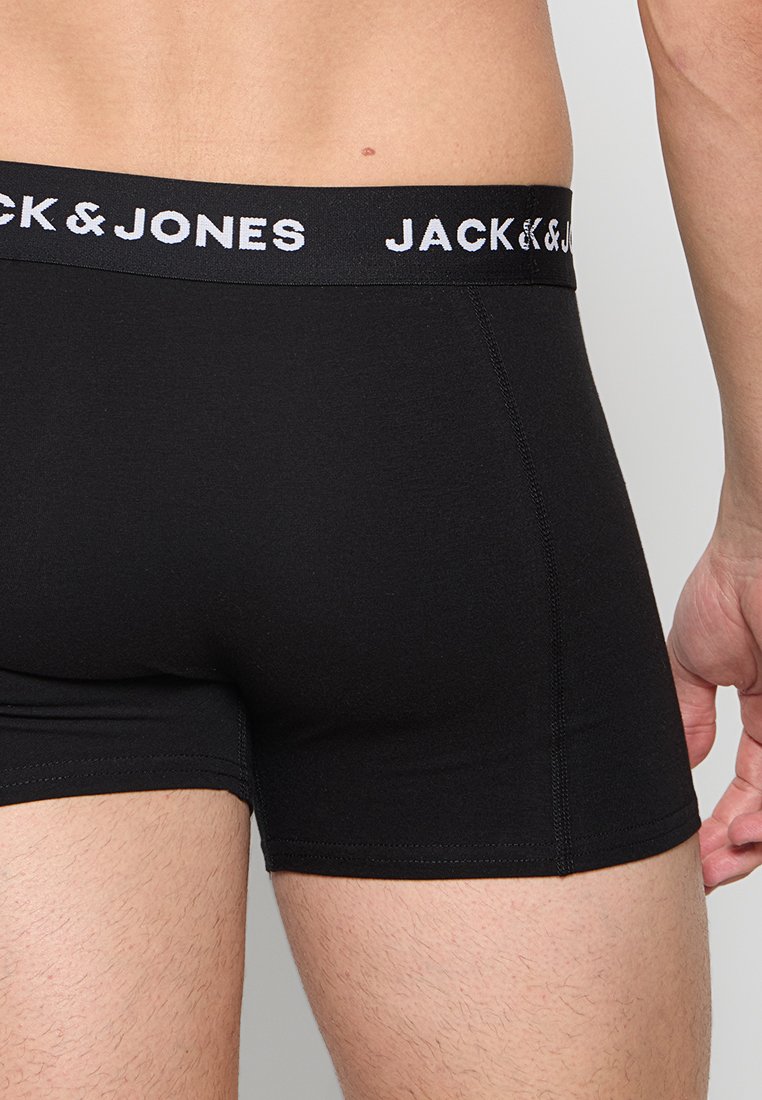 Männlicher Oberkörper und Oberschenkel, die schwarze Jack & Jones Boxershorts tragen, mit sichtbarem schwarzen Logobund auf einfarbigem Hintergrund.