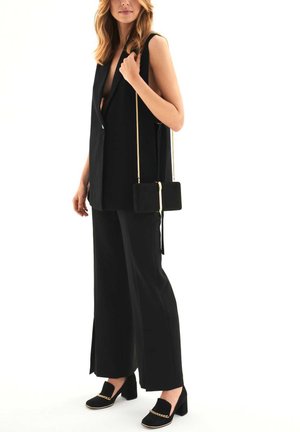 Blazer senza maniche nero con scollatura a V profonda, abbinato a pantaloni a gamba larga e mocassini neri con accento in catena dorata. Porta una piccola borsa nera.