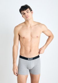 Gray boxer shorts with a black waistband featuring the word "BOSS" in white. The fabric appears smooth and fitted. Model is shirtless.