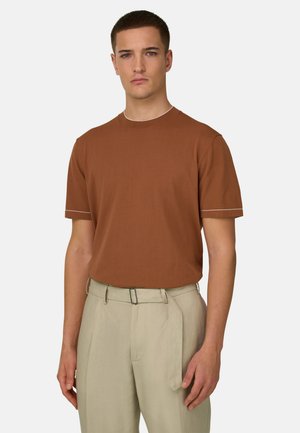 Jeune homme aux cheveux courts portant une chemise marron à manches courtes et un pantalon beige clair taille haute avec une ceinture, debout devant un fond uni.