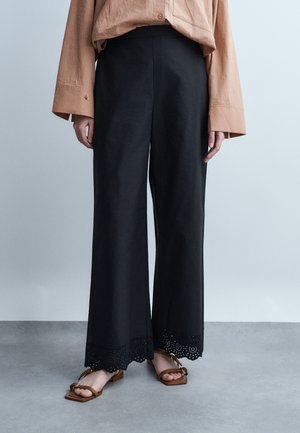 Femme portant un pantalon large noir avec un détail d'œillets à l'ourlet, des sandales à lanières marron et une chemise ample à manches longues de couleur beige, debout sur un sol gris.