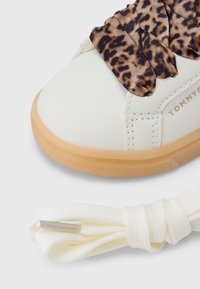 Λευκό αθλητικό παπούτσι με λείο δερμάτινο επάνω μέρος, με επικάλυψη από ύφασμα με λεοπάρ μοτίβο στα κορδόνια και ελαφρώς καφέ λάστιχο σόλας.