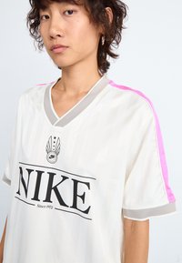 Jersey blanco de Nike con rayas verticales, cuello en V gris, acentos rosas en los hombros y un logo negro en negrita que dice "Desde 1972".