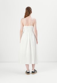 Hvit midi-kjole med tynne justerbare stropper, rynket liv og flytende skjørt. Båret med svarte sneakers med tre hvite striper.