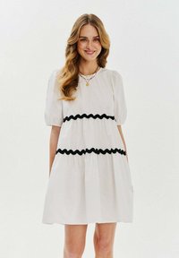 Biele krátke šaty s rukávmi s čiernym ric-rac lemovaním v vrstvenom dizajne. Mäkko látka s voľným strihom, dĺžka po kolená.