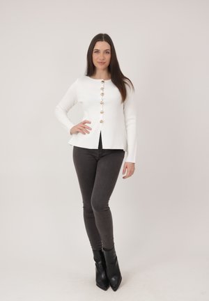 Kobieta z długimi brązowymi włosami, ubrana w białą koszulę na guziki, ciemne obcisłe jeansy i czarne krótkie botki, stojąca z jedną ręką na biodrze.