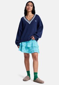 Πουλόβερ σε navy μπλε χρώμα με λαιμόκοψη σε σχήμα V και λευκή ρίγα, φορεμένο πάνω από μια φούστα με βυσσινί και πράσινο καρώ και βολάν. Συνδυάζεται με πράσινες κάλτσες και μπεζ ξύλινα παπούτσια.