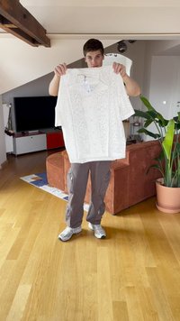 Jaunas vyras, apsirengęs kasdieniais kelnėmis ir sportiniais bateliais, laiko baltą, raštuotą trumpą marškinį patalpoje, fone matyti sofa ir augalas.