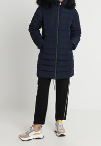 Marinblå puffjacka med en foderhuva i päls, dragkedjeslutning och sidofickor, tillsammans med svarta byxor med vita ränder och klumpiga sneakers.