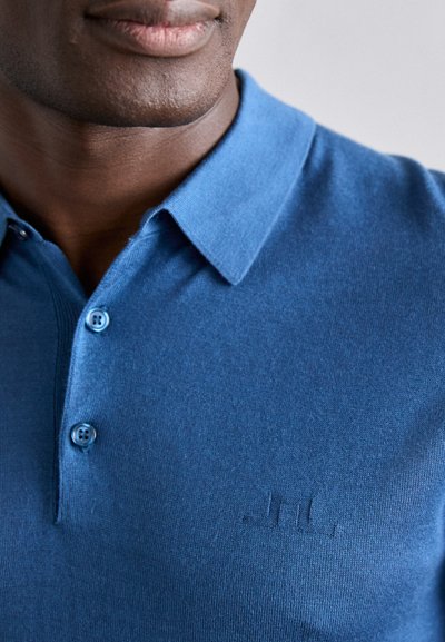 Sinine polo särk pehmest kangast, klassikalise kraega, kahe nupuga ja rinnal reljeefne logo. Sile tekstuur.