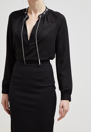 Femme portant un chemisier noir à manches longues avec des cordons à chaînes argentées et une jupe noire ajustée taille haute sur fond clair.