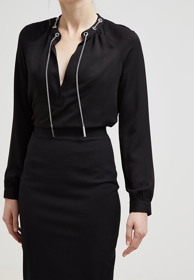 Femme portant un chemisier noir à manches longues avec des cordons à chaînes argentées et une jupe noire ajustée taille haute sur fond clair.