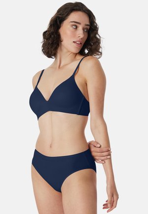 Woman with shoulder-length curly hair wearing a navy blue bra and matching briefs, looking over her left shoulder.