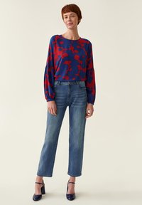 Blusa azul con patrón abstracto rojo, mangas de globo, combinada con jeans rectos de mezclilla azul claro y zapatos de tacón altos en azul marino.
