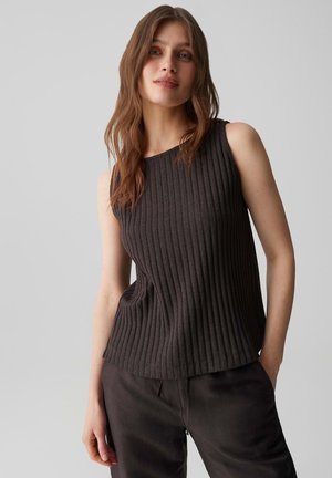 Femme aux longs cheveux bruns portant un haut côtelé sans manches foncé et un pantalon sombre, debout avec une main dans la poche devant un fond uni.