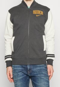 Nike zip-up jack met een donkergrijs lichaam en crèmekleurige mouwen, ribgebreide manchetten met zwart-witte strepen en een gouden logo.