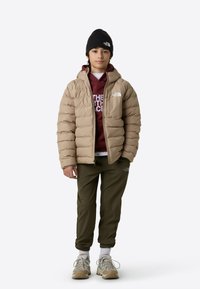 Geacă matlasată bej cu design cusut, purtată peste un hanorac burgund, asortată cu pantaloni jogger olive. Căciulă neagră și adidași bej completează ținuta.