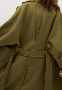 Oliivinvihreä villasekoitetakki, jossa on rento istuvuus, leveät hihat ja vyö vyötäröllä. Takissa on geometrinen ompeleilla koristeltu yksityiskohta.