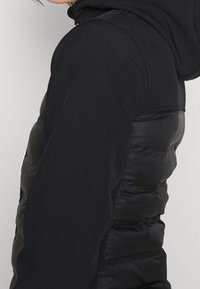 Chaqueta acolchada negra con un exterior suave, que presenta una textura acolchada en el cuerpo y una tela suave en las mangas y la capucha.