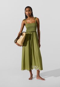 Oliiviroheline varrukateta kleit, millel on veniv ülaosa, voolav seelik ja volangidega ääris. Modell hoiab käsivarrel mustade detailidega õlikiudu kotti.