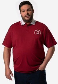 Červené polo tričko s límečkem, který má kontrastní bílé pruhy, logo na levé straně, krátké rukávy a měkkou texturu.