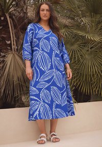 Vestido azul con estampado y diseños de hojas blancas, de longitud media, ajuste relajado y mangas tres cuartos. Combinado con sandalias blancas.