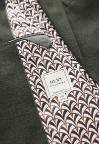 Intérieur de blazer vert texturé avec une doublure blanche arborant un motif géométrique en marron et noir. L'étiquette indique : « NEXT TAILORING, REGULAR FIT ».
