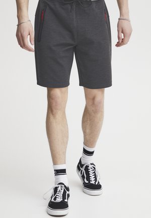 Dunkelgraue Sportshorts mit strukturierter Oberfläche, ausgestattet mit roten Reißverschlusstaschen an den Seiten. Kombiniert mit schwarzen Turnschuhen und weißen gestreiften Socken.