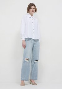 Woman wearing a white button-up blouse, light ripped wide-leg jeans, and gold high-heeled mules, standing against a plain white background.