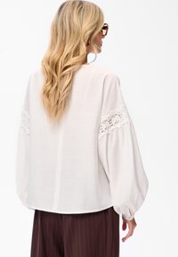 Blondynka z falującymi włosami, nosząca okulary w kratkę tortoise oraz luźną białą bluzkę z koronkowymi detalami na rękawach i brązowe spodnie w prążki.
