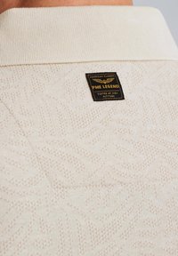Pull en maille texturée couleur crème avec un motif de feuilles subtil, col côtelé et étiquette noire avec des accents dorés. Matière en tricot douce.