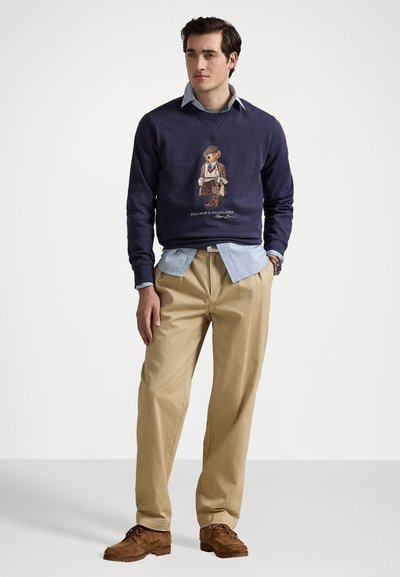 Sweatshirt marine avec un graphisme d'ours, superposé à une chemise à carreaux bleu clair. Un pantalon chino beige et des chaussures en daim marron complètent la tenue.