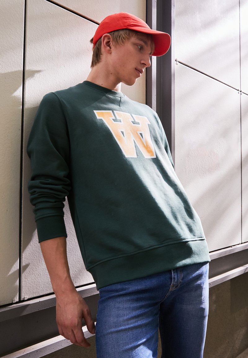 Dunkelgrüner Sweatshirt mit großem cremefarbenem Logo, Rundhalsausschnitt und gerippten Bündchen, kombiniert mit blauen Jeans und einer leuchtend roten Kappe.