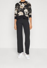 Zwarte trui met bloemenpatroon en ronde halslijn, gecombineerd met zwarte wijde broek en witte sneakers.