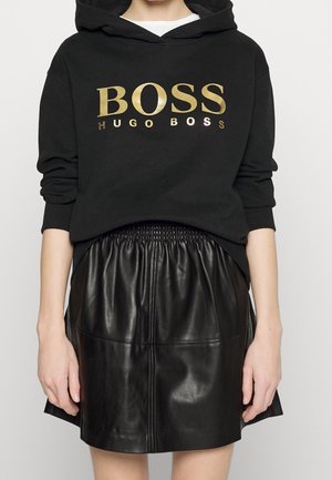 Person iført sort Hugo Boss-hoodie med gyldent tekst "BOSS HUGO BOSS" og sort lædernederdel med rynket talje.