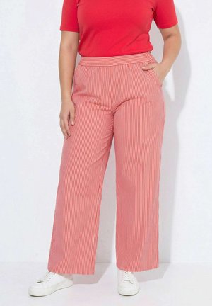 Person iført rød top med korte ærmer og højtaljede, røde og hvide stribede vidde bukser med hvide sneakers, hånd i lommen.