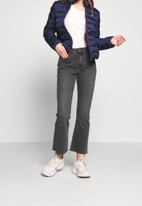 Marinblå pufferjacka med horisontella sömmar, vit t-shirt, grå utställda jeans och ljusa sneakers, som visar en avslappnad outfit.