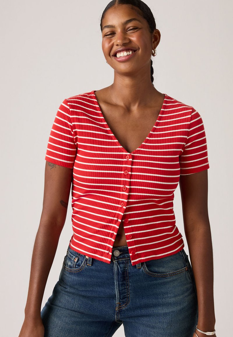 Femme souriante portant un haut à manches courtes à rayures rouges et blanches avec des boutons et un jean bleu, debout devant un fond clair uni.