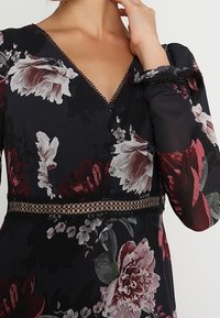 Femme portant une robe noire à fleurs avec un bord en dentelle, un décolleté en V, des manches longues en voile transparent et des boutons décoratifs sur la poitrine.