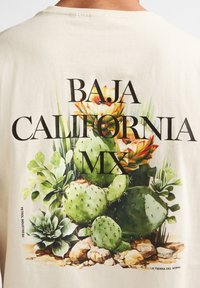 Beiges Baumwoll-T-Shirt mit lebendigem Kaktus- und Blumenmuster auf der Rückseite, mit schwarzem Text "BAJA CALIFORNIA MX."