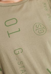 Olive-green fabric with green block letters and partial text "G-STAR RAW" printed on a shirt's chest area.