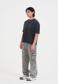 Sort oversized bomulds t-shirt med broderet tekst, sammen med grå denim bukser med hvide grafiske mønstre og cargo-lommer. Hvide sneakers.