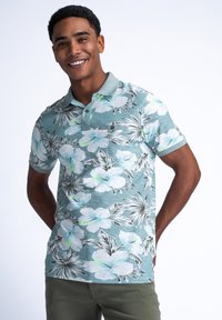 Teal värinen lyhythihainen polo, jossa on valkoisia ja vaaleansinisiä hibiskikukkia sisältävä kukkakuosi, yhdistettynä oliivinvihreisiin housuihin.