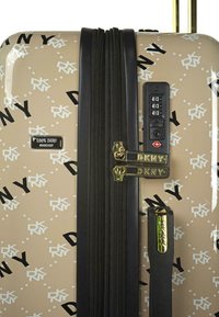 Maleta de hard-shell en beige con un patrón de logo de DKNY en blanco y negro. Cuenta con dobles cremalleras, cerradura de combinación y acentos dorados.