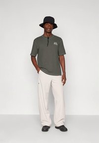 Hombre con sombrero de cubo negro, camisa de manga corta verde oscuro, pantalones anchos beige claros y zapatos negros, de pie contra un fondo liso.