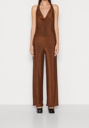 Kvinde iført en ærmeløs, brun skinnende halterneck-jumpsuit med vide bukseben og sorte flade sko, stående mod en ensfarvet baggrund.