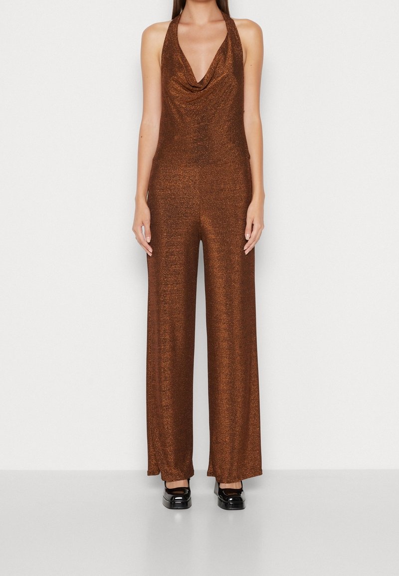 Femme portant une combinaison sans manches marron scintillante à col halter avec pantalon à jambes larges et chaussures plates noires, debout devant un fond uni.