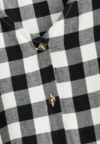 Tissu à carreaux noir et blanc avec un détail de bouton. Présente un design en v et un motif de couture visible. Matière en coton.