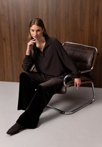 Jeune femme en tenue sombre ample assise sur une chaise moderne en cuir et chrome contre un mur en panneaux de bois, la main reposant près du visage.
