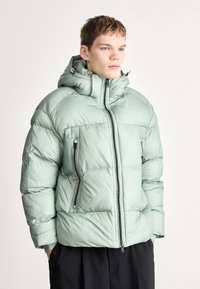 Απαλό πράσινο μπουφάν puffer με κουκούλα, διαθέτει εμπρόσθιο φερμουάρ, τσέπες με φερμουάρ στα πλάγια και καπιτονέ υφή.